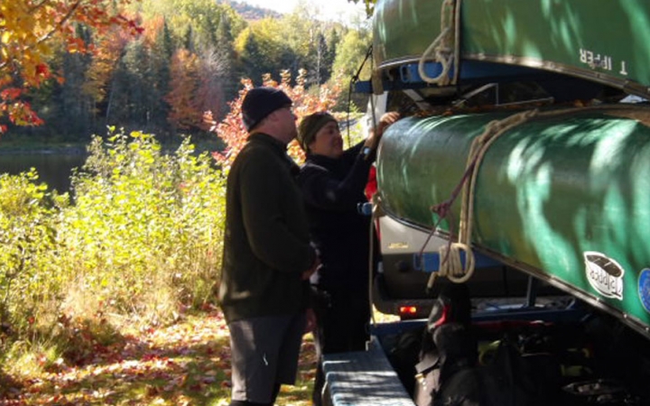 Two people examine canoes stacked on a trailer. Behind them, you can see fall foliage. 